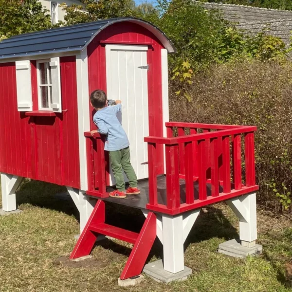 Kind steht vor rotem Bauwagen aus Holz im Garten – Der Kleine Bauwagen als Spielhaus